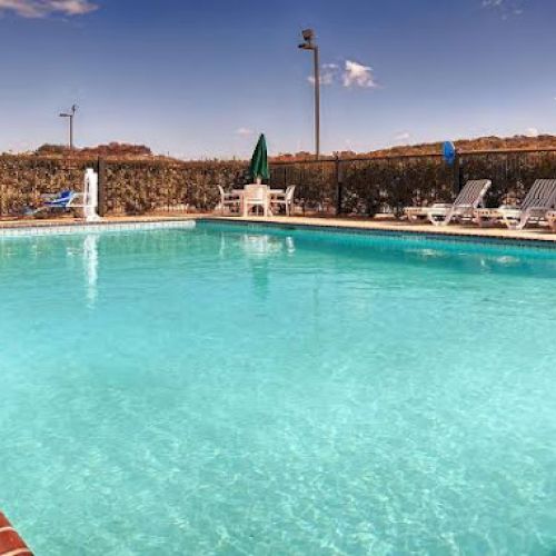 A clear swimming pool surrounded by lounge chairs and a sunny sky. The pool is bordered with bricks and a fence runs along the perimeter.