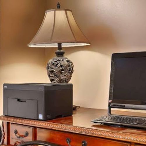 A desk with a computer, printer, lamp, and office chair near window blinds.