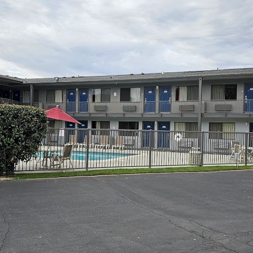 The image shows a two-story motel with a pool area, surrounded by a fence and umbrellas, and an asphalt parking area in front.