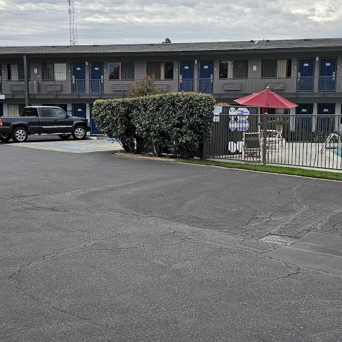 The image shows a two-story motel with a parking lot, a pool area with lounge chairs, and a red umbrella on a cloudy day.