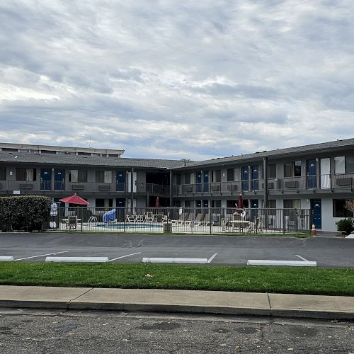 The image shows a two-story motel with a swimming pool in the center, surrounded by a parking lot under a cloudy sky.