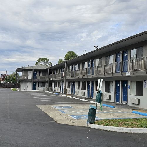 The image shows a two-story motel with a parking lot, featuring blue doors and reserved accessible parking spaces.