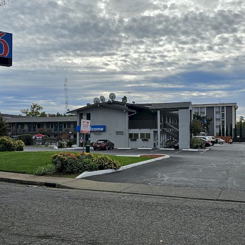 The image shows a roadside motel with a prominent sign and parking lot under a cloudy sky, featuring a palm tree.