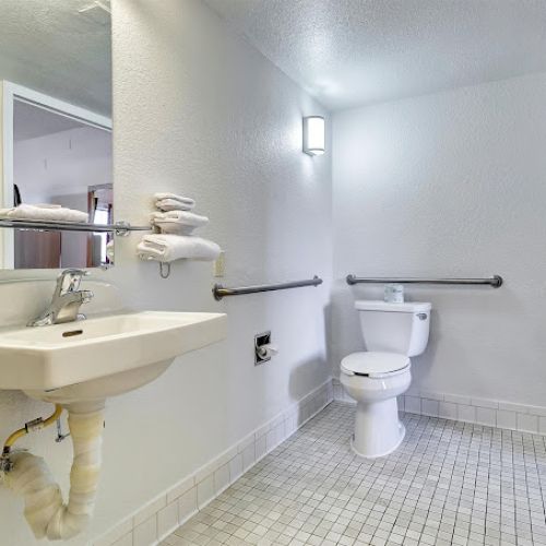 A bathroom with a wall-mounted sink, mirror, toilet, grab bars, and white tiled floor is shown.