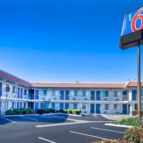 The image shows a Motel 6 building with a two-story structure, empty parking lot, and a tall sign displaying the number 6, under a clear blue sky.