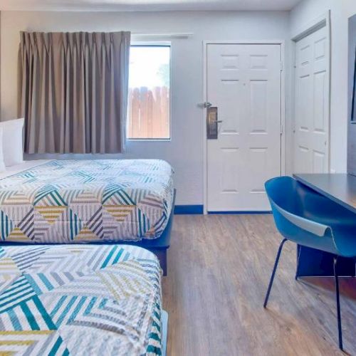 A modern hotel room with two beds, a desk with a lamp, a chair, TV, and a window with curtains.