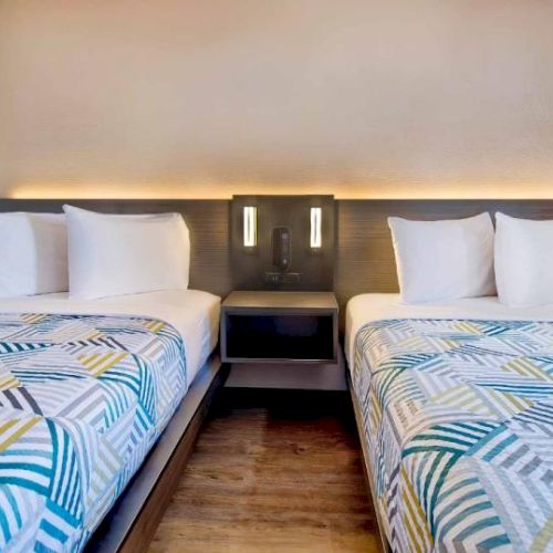 The image shows a hotel room with two single beds, patterned bedspreads, a nightstand in between, and wall lighting.