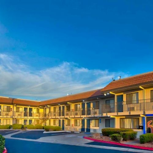 The image shows a Motel 6 building with an elevated sign, two-story layout, and parking in front, under a clear blue sky.