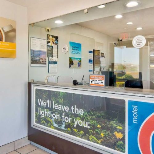 The image shows a Motel 6 reception area with signs, brochures, and a counter with a protective screen. The slogan 