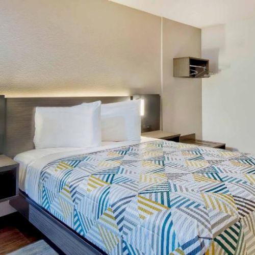 The image shows a modern hotel room with a neatly made double bed, geometric patterned bedding, bedside tables, and a small shelf on the wall.