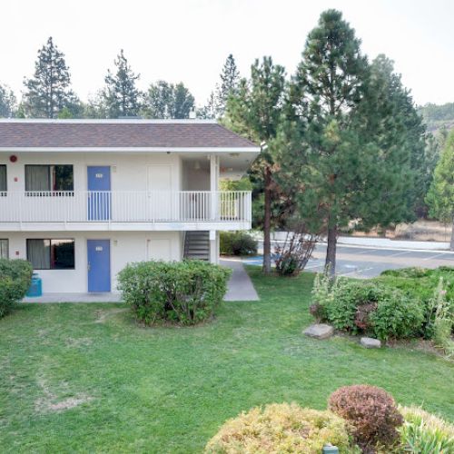 A two-story building with blue doors is surrounded by trees and shrubs, with a road visible in the background.