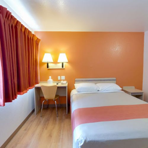 A simple hotel room with a bed, desk, chair, lamp, and window with curtains. The walls are painted orange and white.