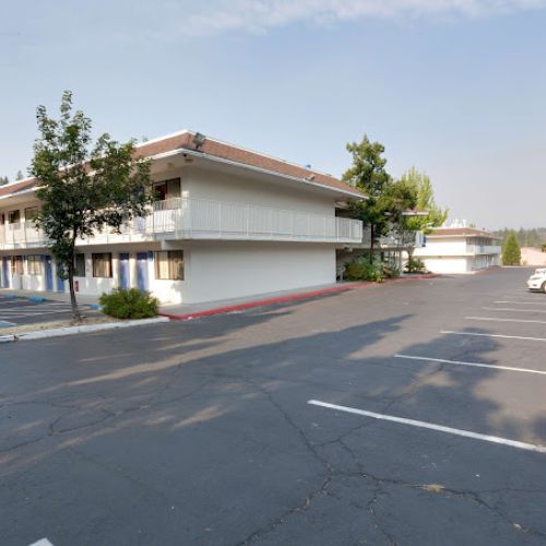 A two-story motel with parked cars and trees around it, featuring a clear, sunny day in the background.