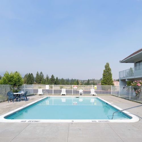 A fenced outdoor swimming pool with lounge chairs and a seated area next to a building, surrounded by trees under a clear sky.