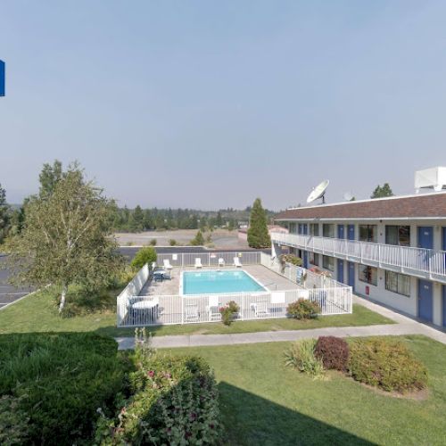 The image shows a two-story motel with a pool, surrounded by greenery, and a Motel 6 sign towering in the background.