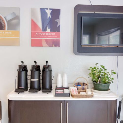 Coffee station with dispensers, cups, and condiments beneath wall art and a TV, flanked by chairs and a plant on the counter.