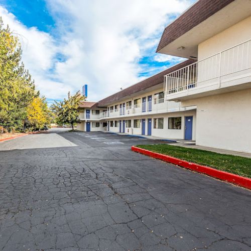 A two-story motel building with blue doors, surrounded by trees and a cracked asphalt parking lot under a partly cloudy sky.