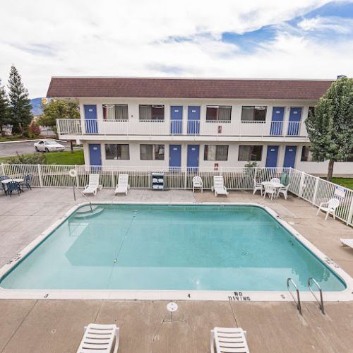 A motel with a blue and white exterior has a fenced swimming pool and several lounge chairs surrounding it.