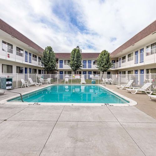 An outdoor swimming pool surrounded by lounge chairs, located within a two-story motel courtyard with trees and a partly cloudy sky.