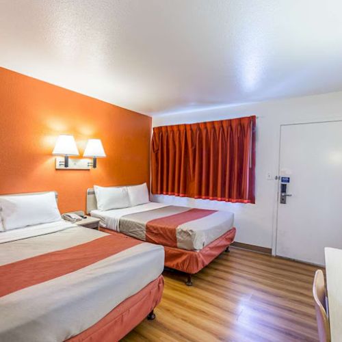 The image shows a hotel room with two beds, orange accents, wooden flooring, desk, chair, and wall lighting under a bright ceiling.