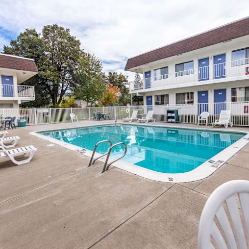 An outdoor swimming pool surrounded by lounge chairs and white fences, with two motel buildings in the background under a cloudy sky.