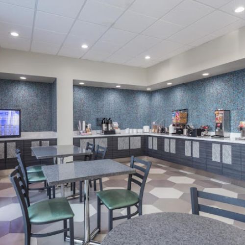 A modern dining area with tables, chairs, a refrigerator, coffee machine, and beverage dispenser on the counter against a patterned wall.