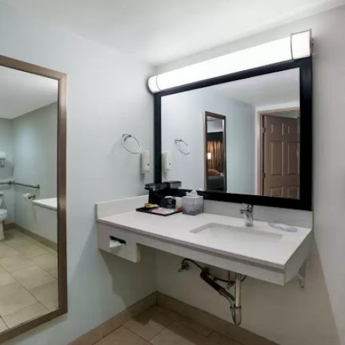 The image shows a bathroom with a large mirror, sink, wall-mounted hairdryer, and a doorway leading to a toilet area.