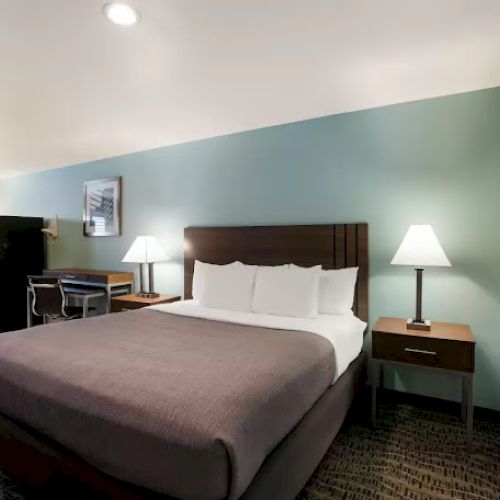 The image shows a hotel room with a bed, bedside lamps, artwork, a chair, and a small fridge against a soft blue wall.