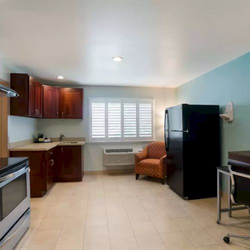 A kitchen area with an oven, wooden cabinets, a refrigerator, an armchair, a desk, and a chair. The room has tiled floors and a window.