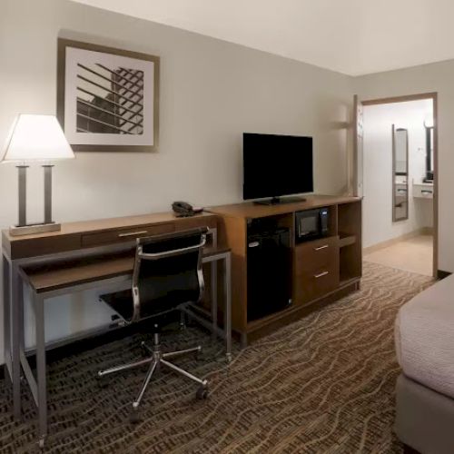 A hotel room features a desk, chair, TV, lamp, artwork on the wall, and a bed, with a doorway leading to another room or area.