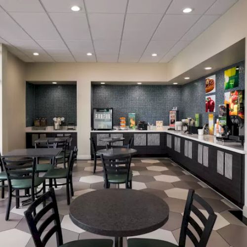 A small dining area with tables and chairs, next to a self-service counter offering drinks and snacks on a tiled floor.