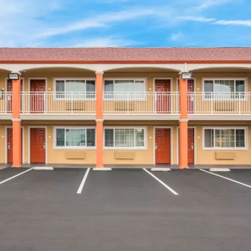 The image shows a two-story motel with orange and beige exterior, a staircase, and a parking lot with empty spaces.