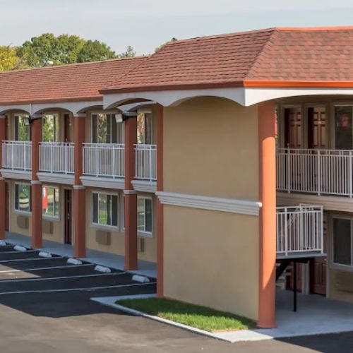 The image shows a two-story motel with a red-tiled roof, exterior corridors, and white railings on a sunny day.