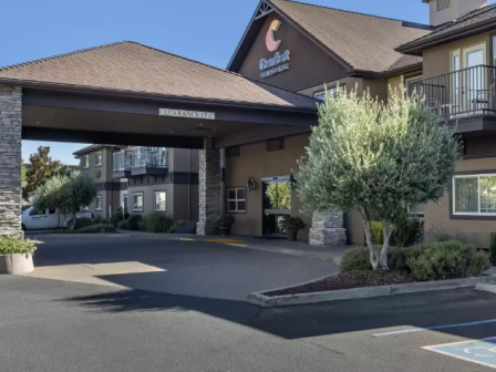 This image shows the entrance of a hotel with a covered driveway, trees, and a visible logo on the building.