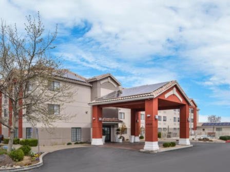 The image shows a hotel entrance with a covered driveway, red columns, and a landscaped area, under a partly cloudy sky.