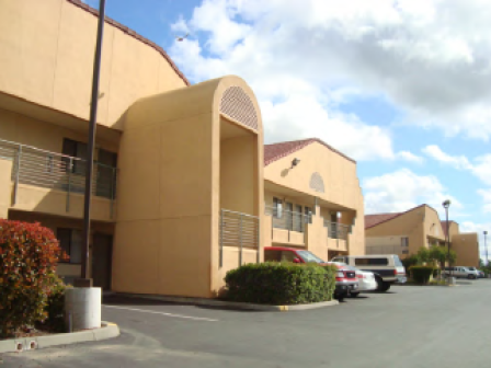 The image shows a motel or hotel building with a parking lot and several cars parked outside under a partly cloudy sky.