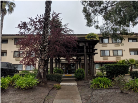 The image shows the entrance of a three-story building with surrounding trees, plants, and a pathway leading to the door.