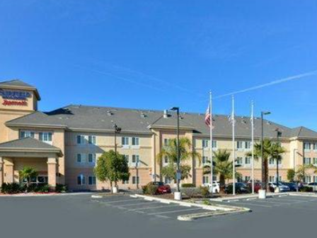 The image shows a Fairfield Inn & Suites hotel with a parking lot and several palm trees under a clear blue sky.