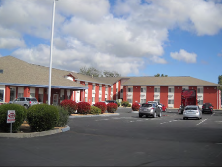 A red two-story motel with a parking lot and several cars, surrounded by shrubs, under a partly cloudy sky.