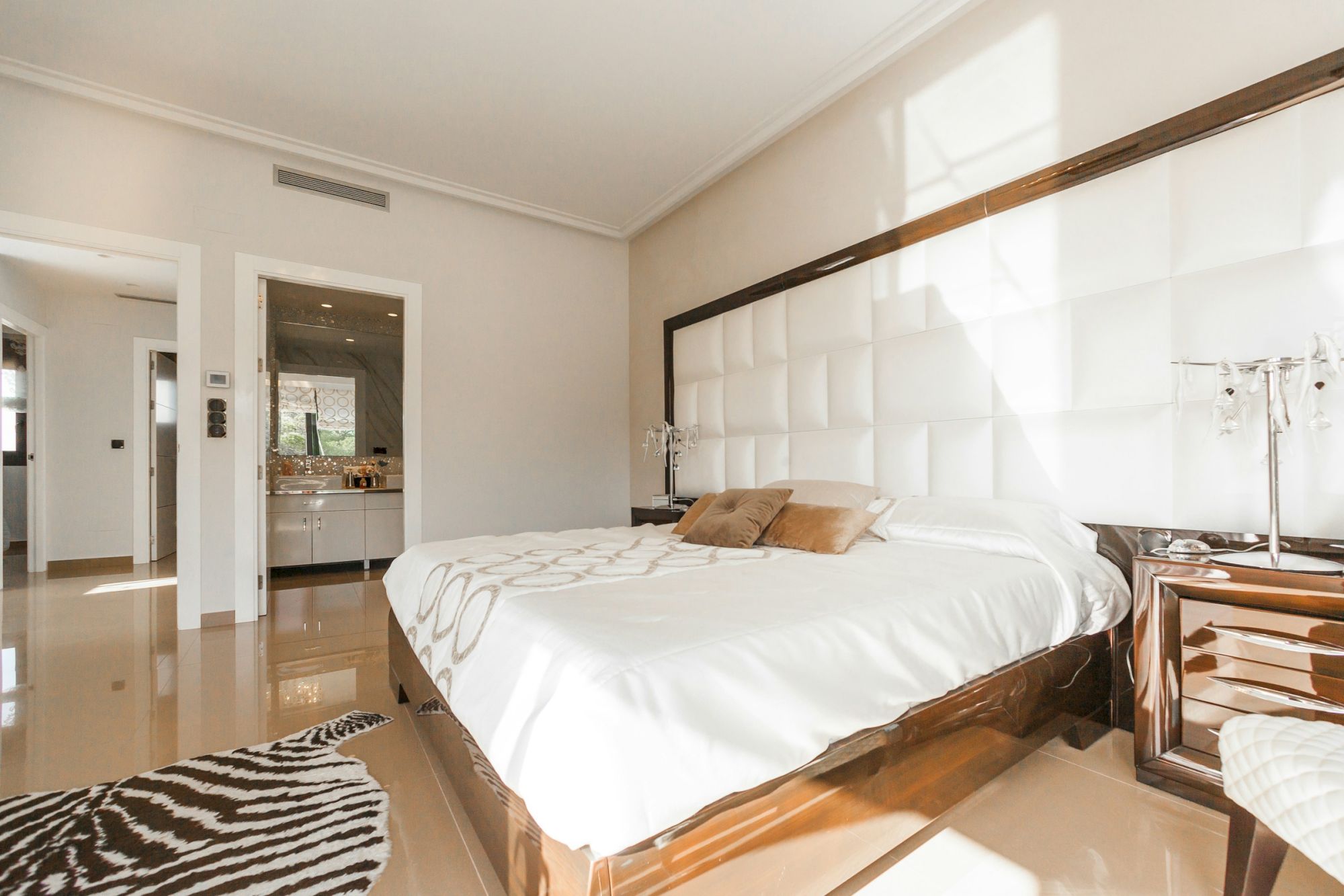 The image shows a modern bedroom with a large bed, a decorative rug, and a view into a bathroom. The space is bright and elegant.