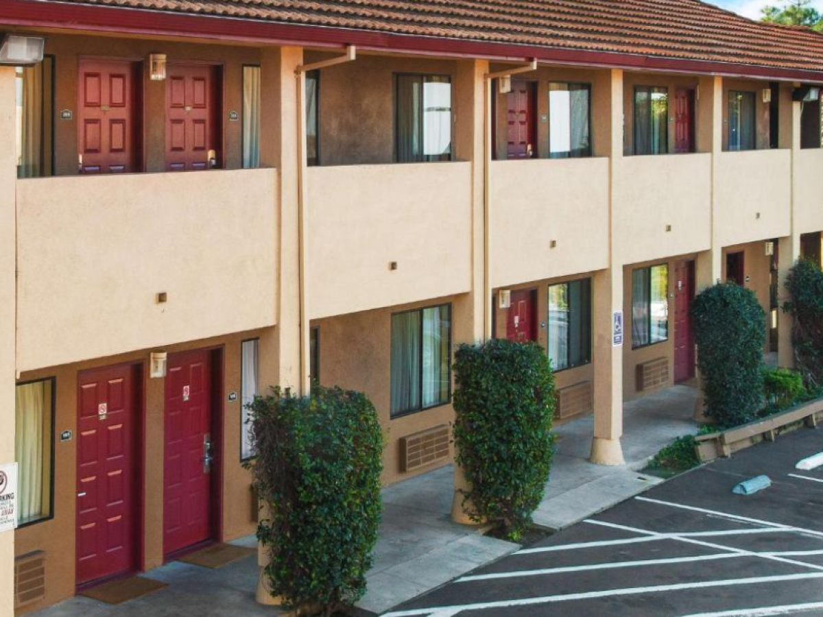 A two-story motel with red doors, beige walls, and trimmed bushes along the walkway, featuring a parking area with marked spaces.