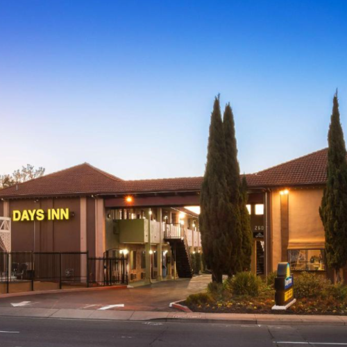 The image shows a Days Inn hotel with a tiled roof, surrounded by trees, under a clear blue sky at dusk.