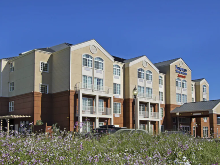 A multi-story hotel building with a sign, surrounded by a green lawn with flowers under a clear blue sky.