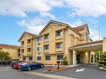 The image shows a mid-sized hotel with a beige exterior, a few parked cars, and a covered entrance, set under a partly cloudy sky.