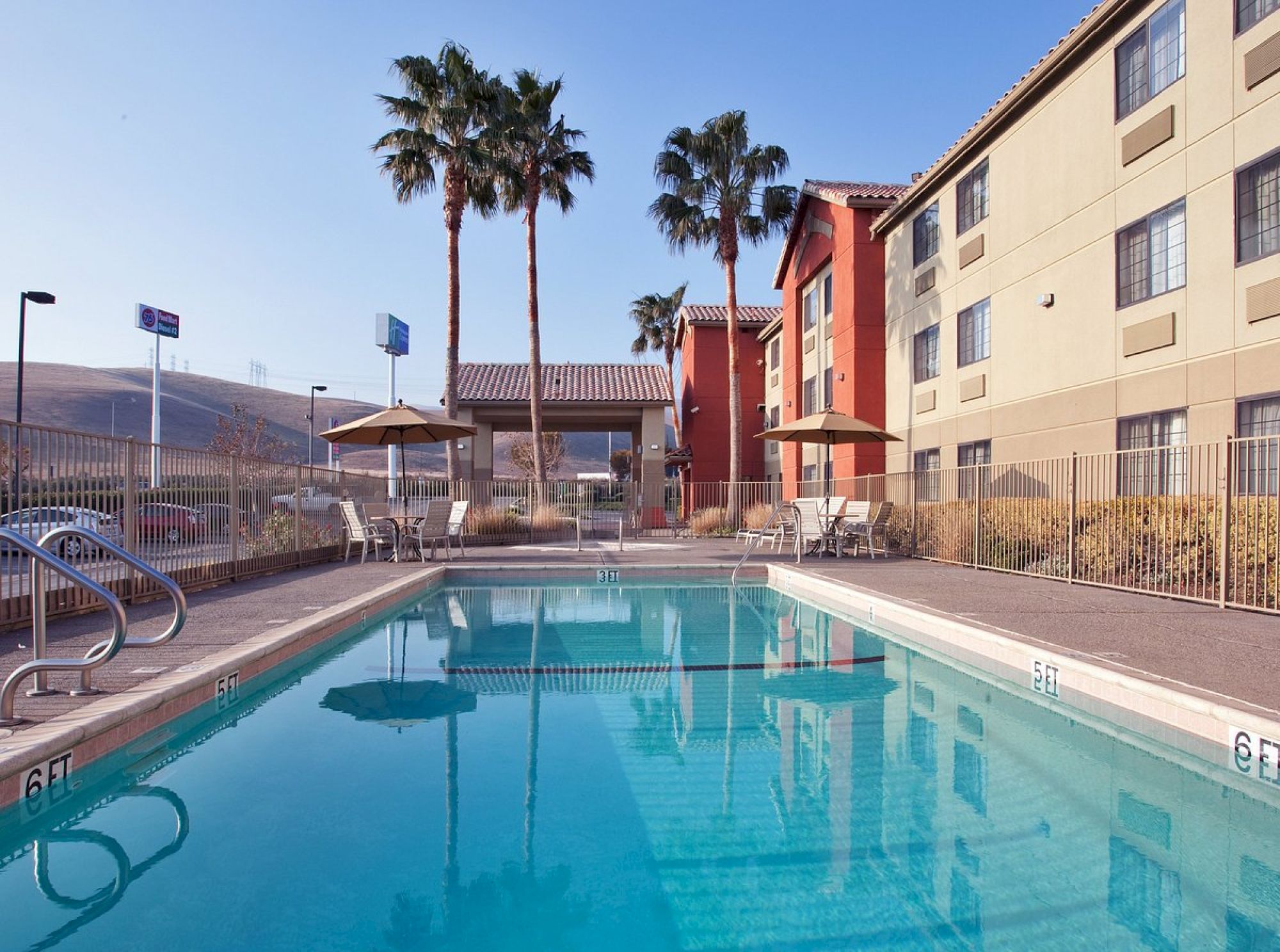 A hotel pool area with clear water, surrounded by lounge chairs and palm trees, adjacent to a multi-story building under a clear sky.