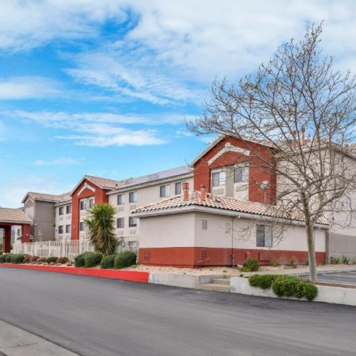 The image shows a two-story hotel building with a red roof, surrounded by a parking lot and minimal landscape under a partly cloudy sky.