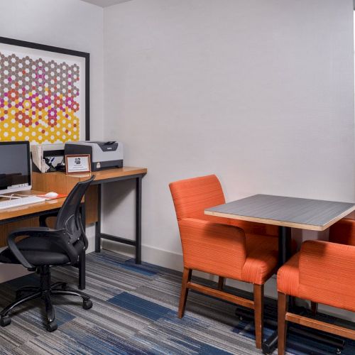 The image shows an office with a computer, printer, art on the wall, a desk with a chair, and a small table with two orange chairs.