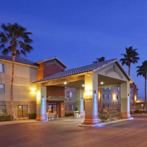 A hotel entrance is lit in the evening, featuring palm trees and a three-story building under a blue sky.