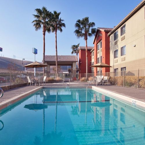 An outdoor swimming pool with clear water, surrounded by a hotel building, lounge chairs, umbrellas, and palm trees under a clear sky.