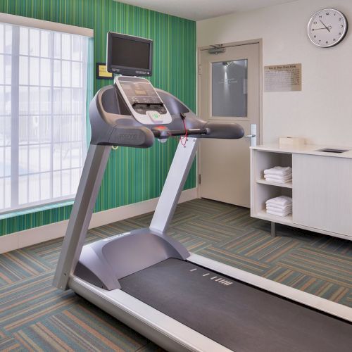 The image shows a gym room with a treadmill, TV, clock, and a shelf with towels and cups, set against a green-striped wall.